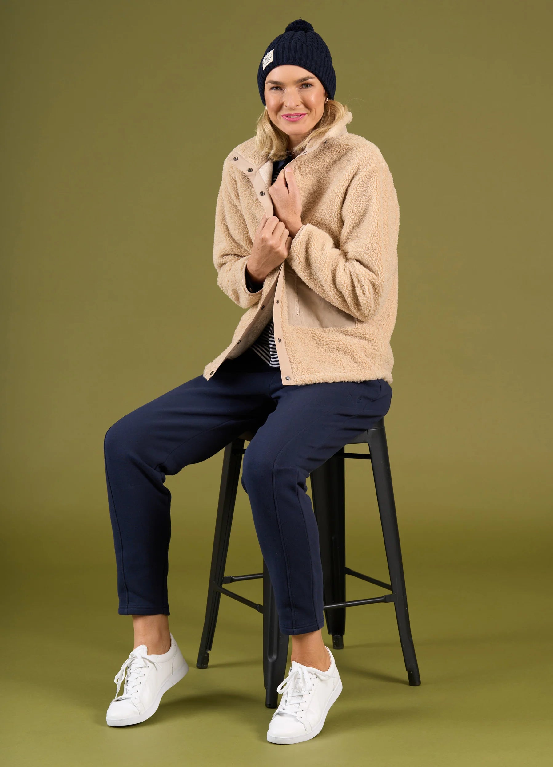 Person wearing a beige jacket, navy pants, and white sneakers sitting on a black stool against a green background.