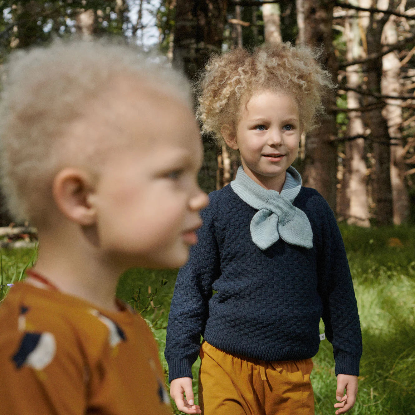 Two children standing in a forested area, one wearing a navy sweater and yellow pants, the other in an orange shirt with bird designs.