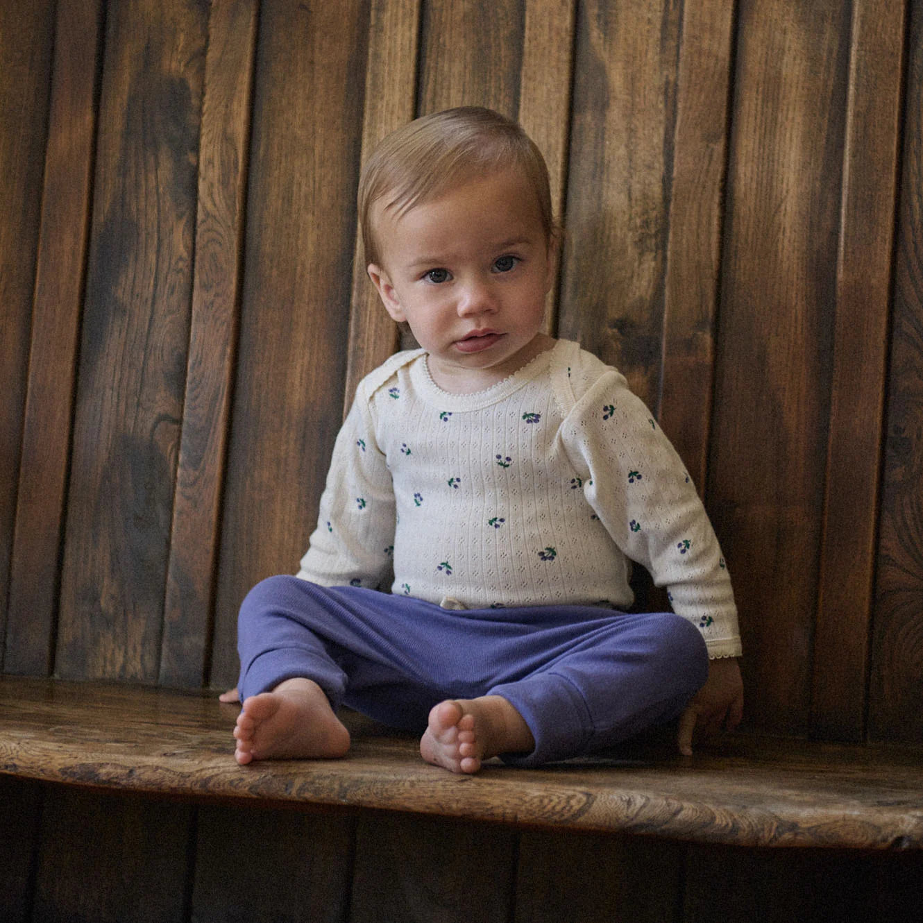 Baby sitting on a wooden bench wearing a white sweater with blue patterns and purple pants.