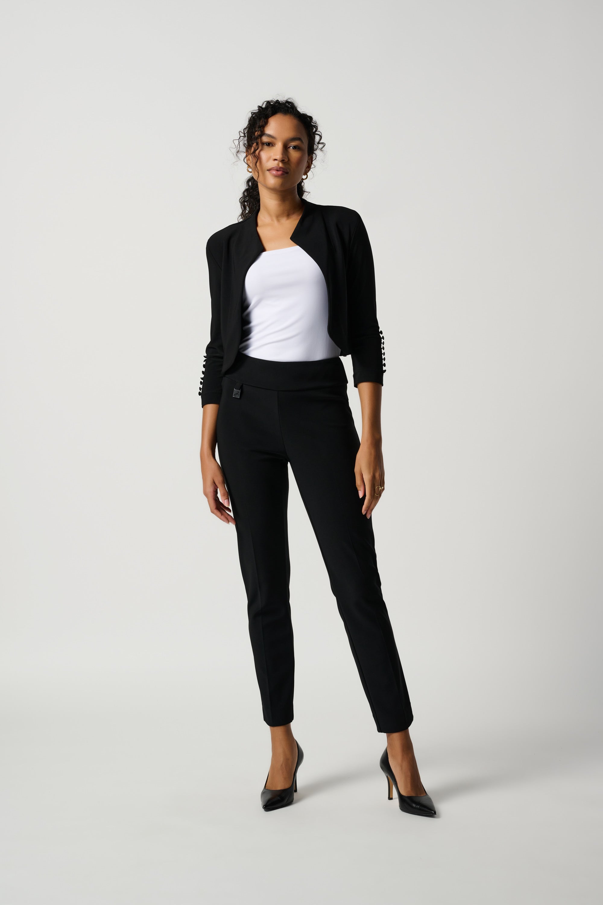 A woman standing in a studio setting, wearing a white top and black tailored slim pants with a subtle ornament.