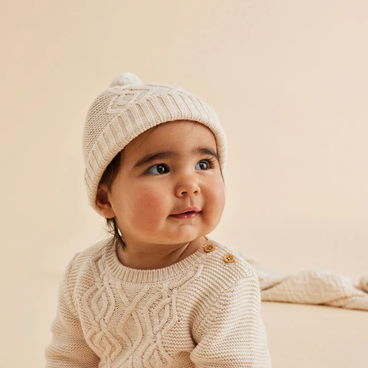 Baby wearing a beige knit sweater and hat against a beige background