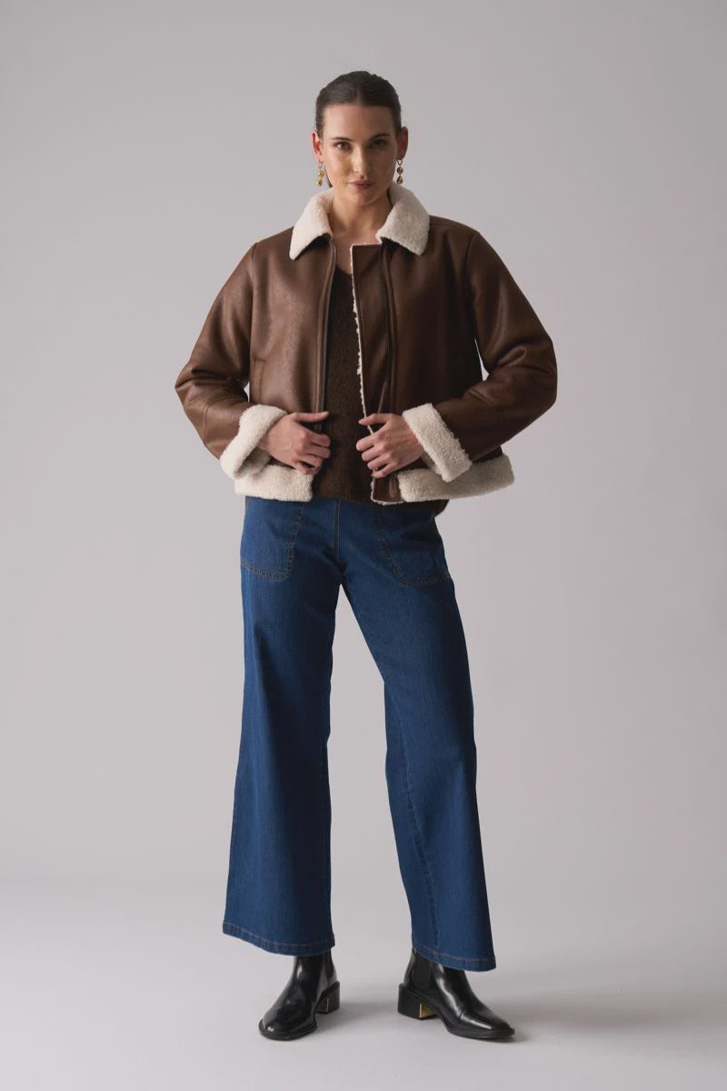 Person wearing a brown shearling jacket with white fur trim, blue jeans, and black shoes on a gray background