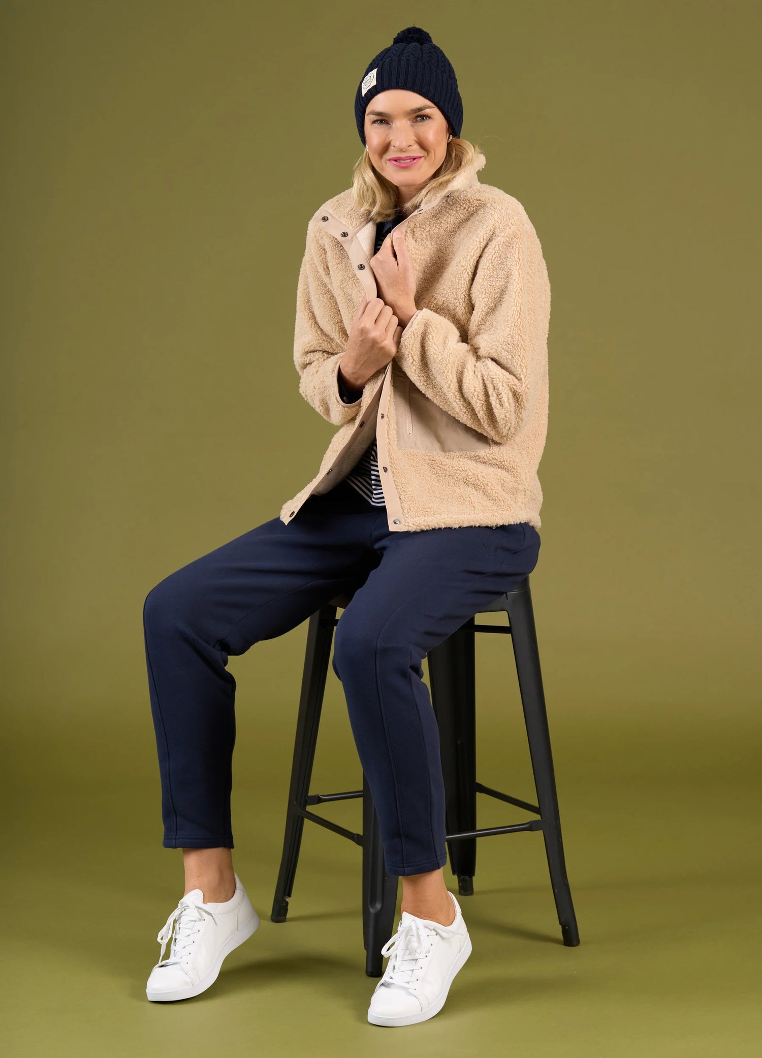 Person wearing a beige jacket, navy pants, and white sneakers sitting on a black stool against a green background.
