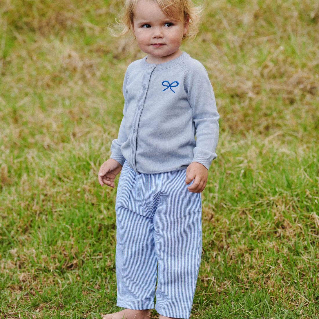 Child wearing a light blue shirt with a bow design and striped pants standing in a grassy field.