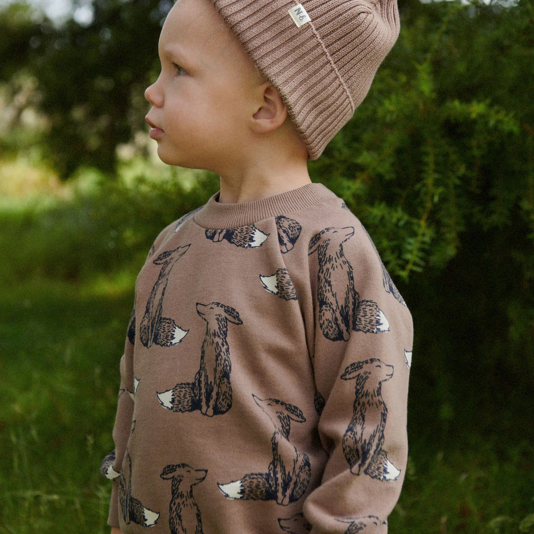 Child wearing a brown sweater with animal patterns and a matching beanie outdoors.