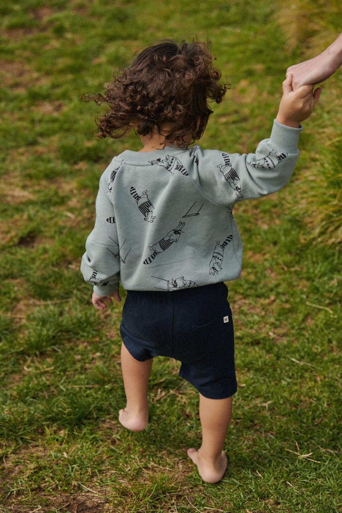 Child wearing a light green hoodie with black patterns and navy shorts standing on grass.