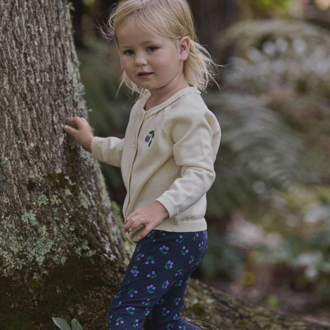 Child in a forest setting wearing a cream sweater and dark patterned pants.