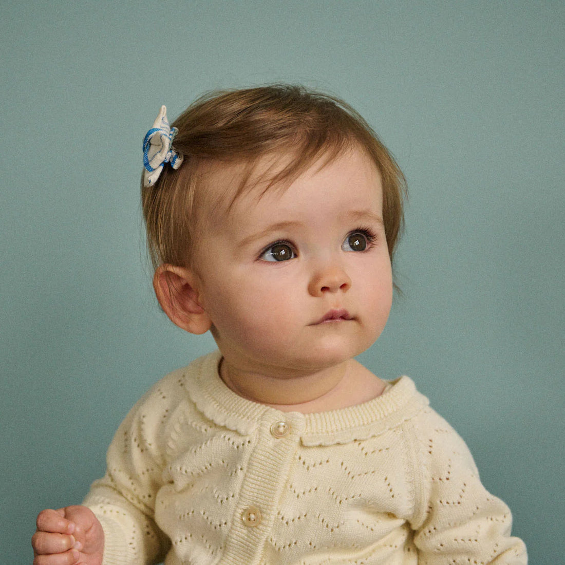 Baby wearing a cream sweater with a blue and white hair accessory against a teal background