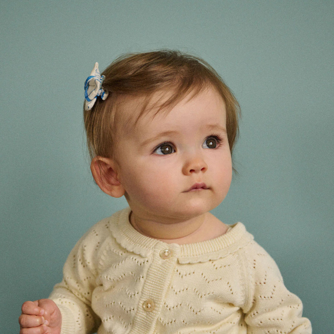 Baby wearing a cream sweater with a blue and white hair accessory against a teal background