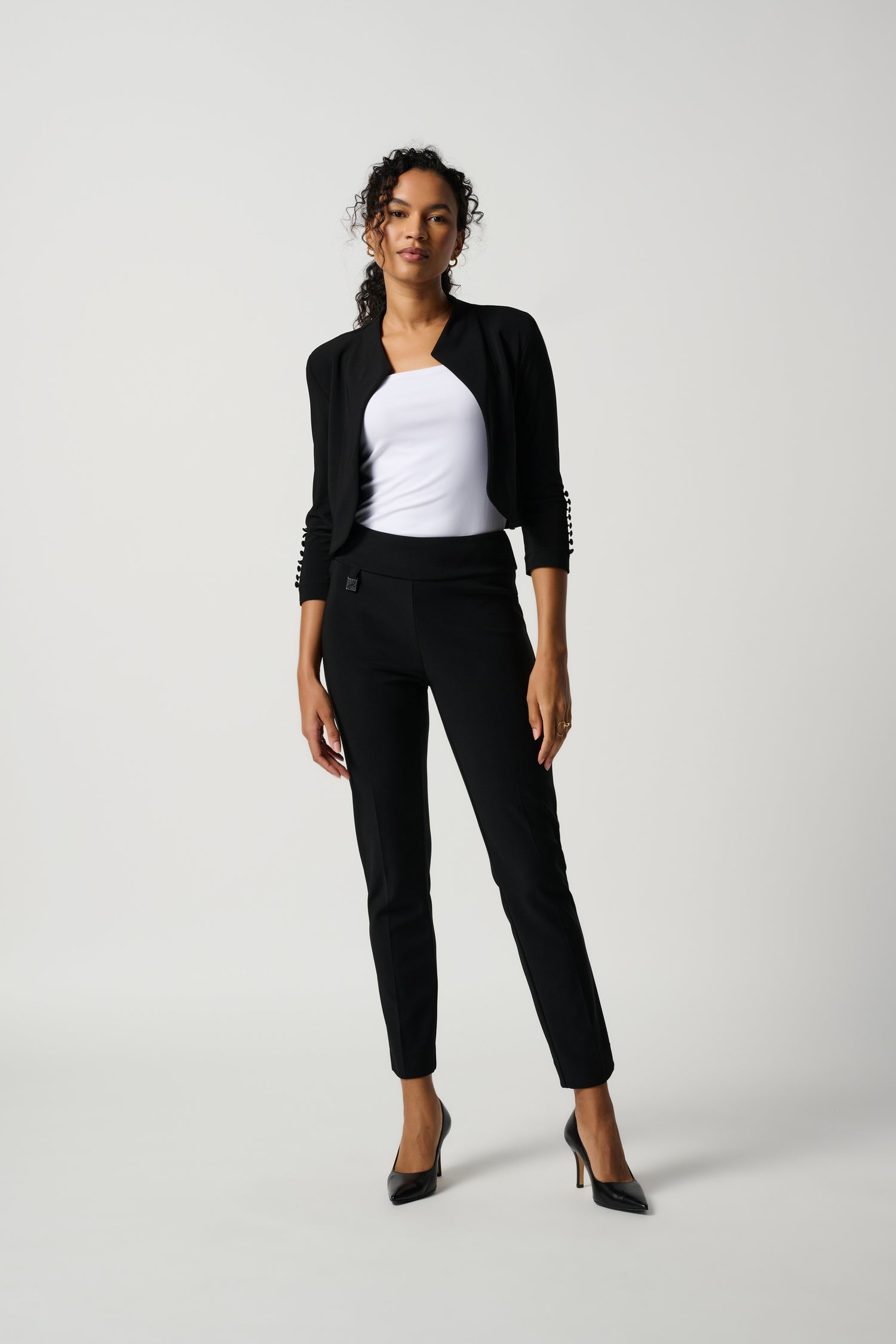 A woman standing in a studio setting, wearing a white top and black tailored slim pants with a subtle ornament.