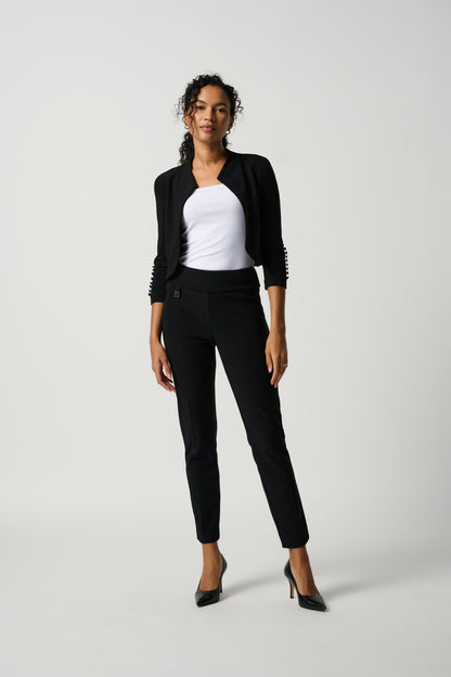 A woman standing in a studio setting, wearing a white top and black tailored slim pants with a subtle ornament.