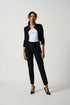 A woman standing in a studio setting, wearing a white top and black tailored slim pants with a subtle ornament.