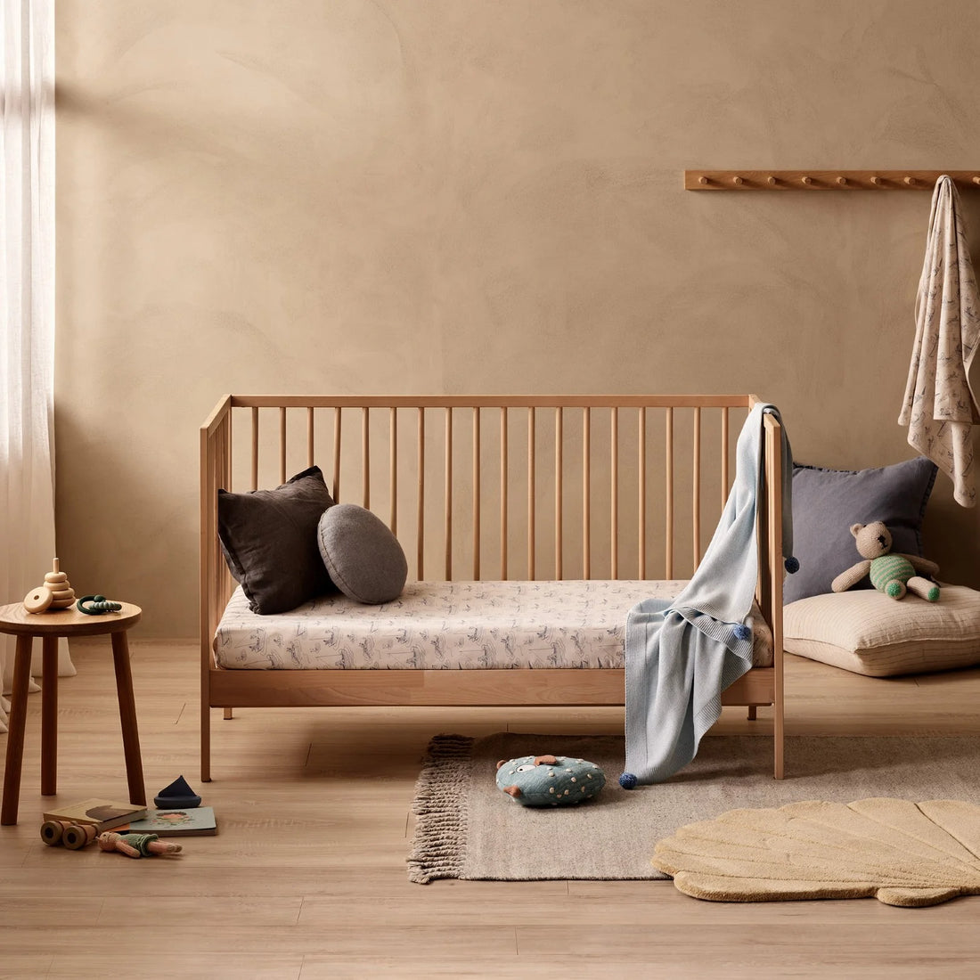 Wooden crib with bedding and toys in a room with a beige wall and wooden floor.